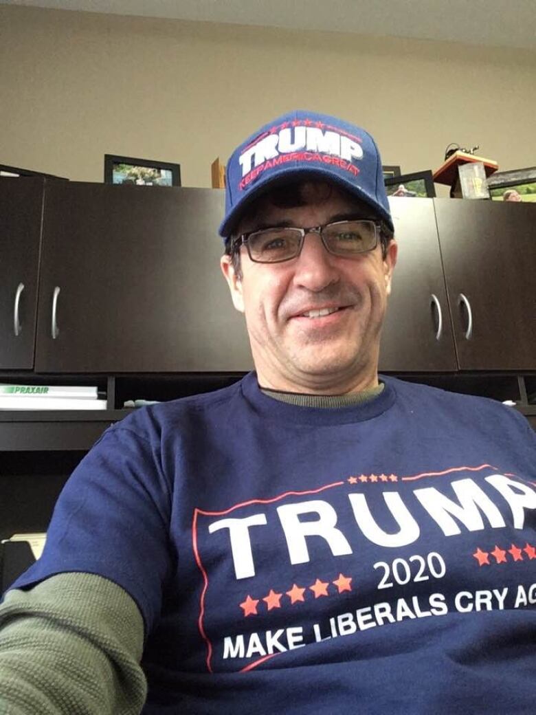 A man wearing glasses smiles at the camera. He is also wearing a blue baseball cap that says 