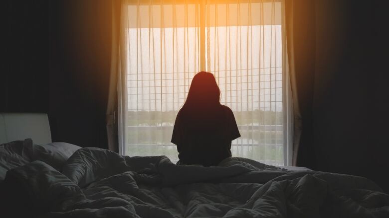 A stock photo shows the back of a woman silhouetted against a window as she sits on a bed in a hotel room.