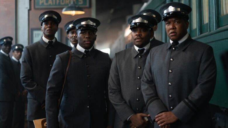 Six men stand in train porter uniforms with hats, in front of train.