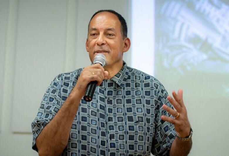 A Black man wearing a patterned short sleeve button up. He is holding a mic 