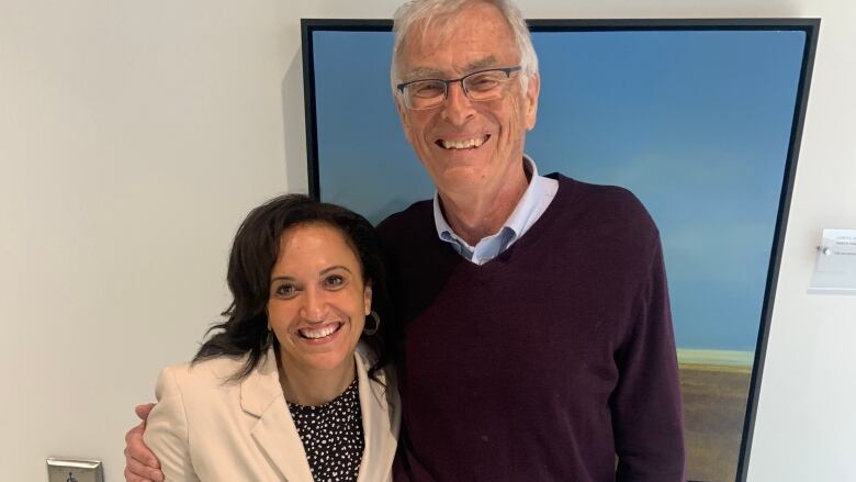 A Black woman is standing beside a white man. She is wearing a cream blazer. He is wearing a navy sweater