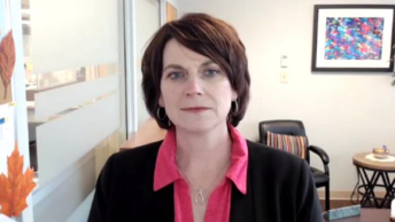 A woman with brown hair wears a dark blazer and a pink shirt while sitting in her office.