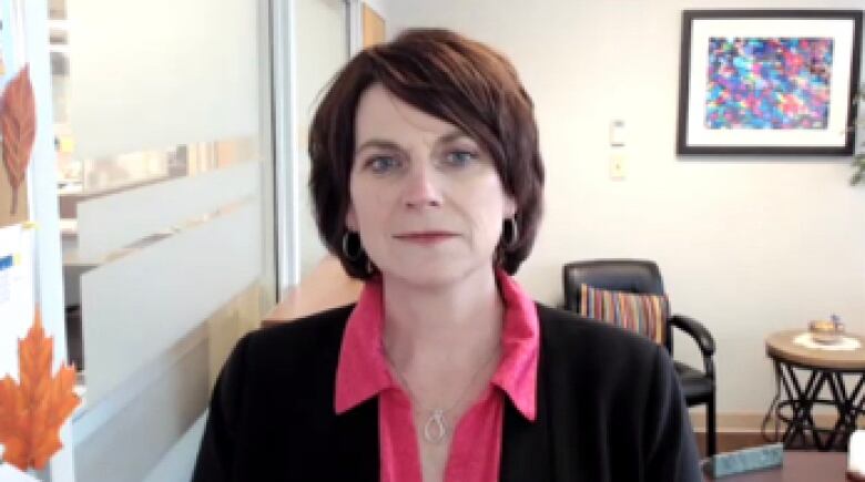 A woman with brown hair wears a dark blazer and a pink shirt while sitting in her office.