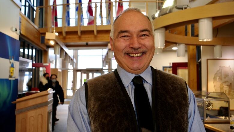 A balding man wearing a shirt and tie draped by a fur vest