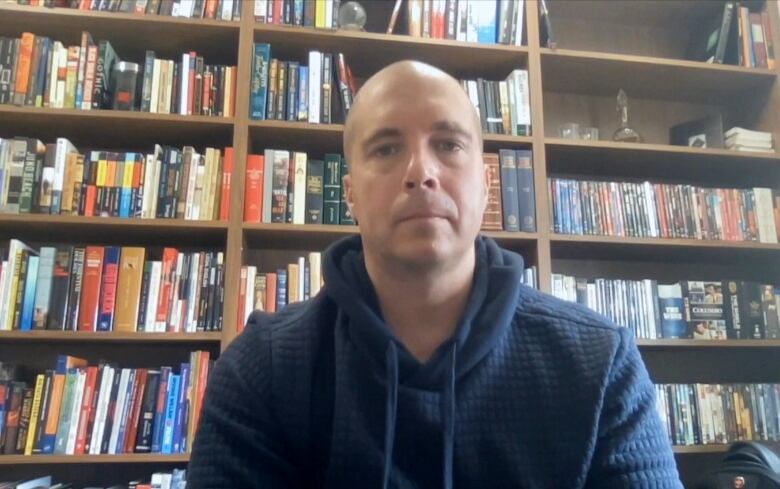 A bald man wearing a blue hooded sweater sits in front of a bookshelf.