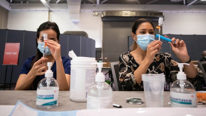 Two women hold syringes over small bottles at a table with plastic cups, hand sanitizer in a vaccine clinic