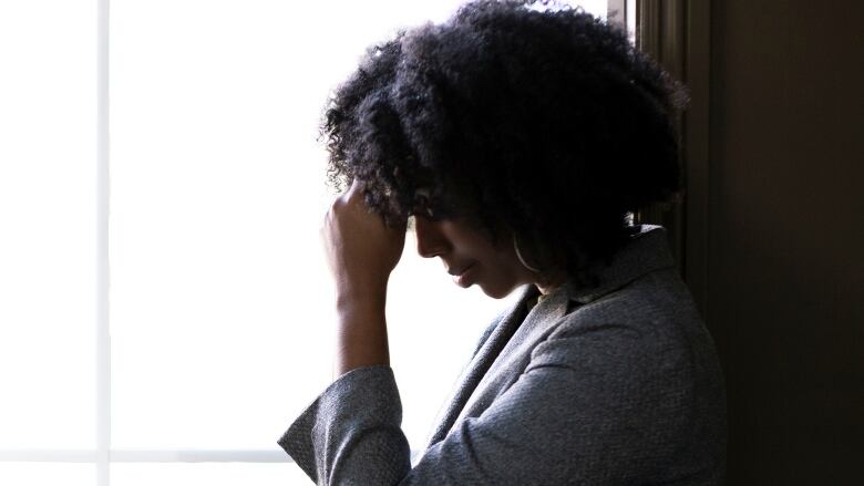 A Black woman stands in front of a window with a hand against her forehead, 