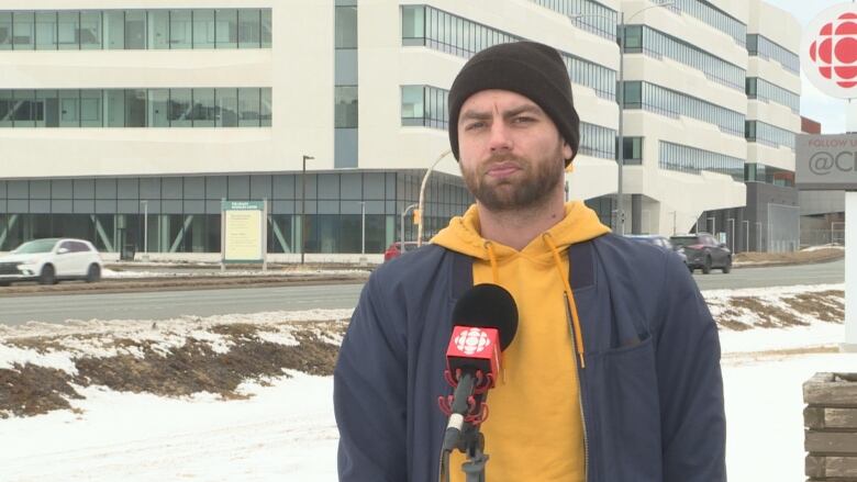 A young man in his 30s with a beard and a yellow hoodie, wearing a beanie. He's standing outdoors.