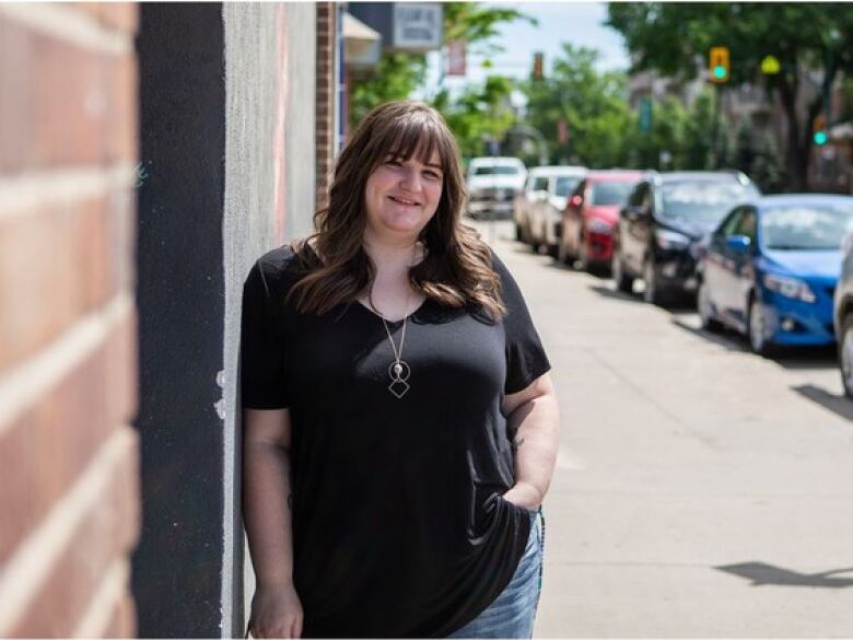 A smiling person with long hair and wearing a black T-shirt, a long silver necklace and blue jeans leans against a brick wall on a sunny city street.