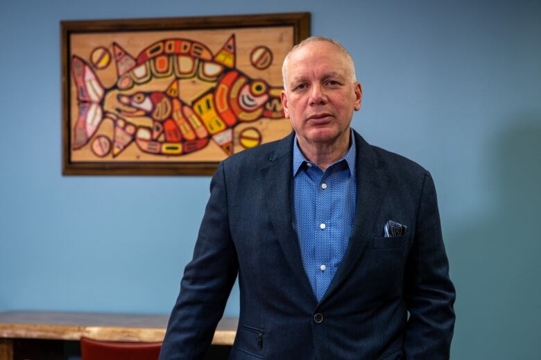 A man with thinning white hair in a dark blue suit and blue shirt stands in front of an Indigenous painting. 