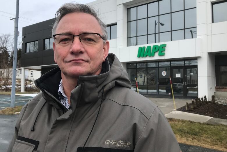 A man looks straight ahead. He stands in front of a building with the letters NAPE above the front doors.