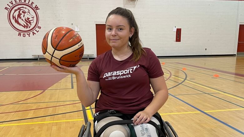 A woman in a wheelchair with brown hair in a ponytail holds up a basketball