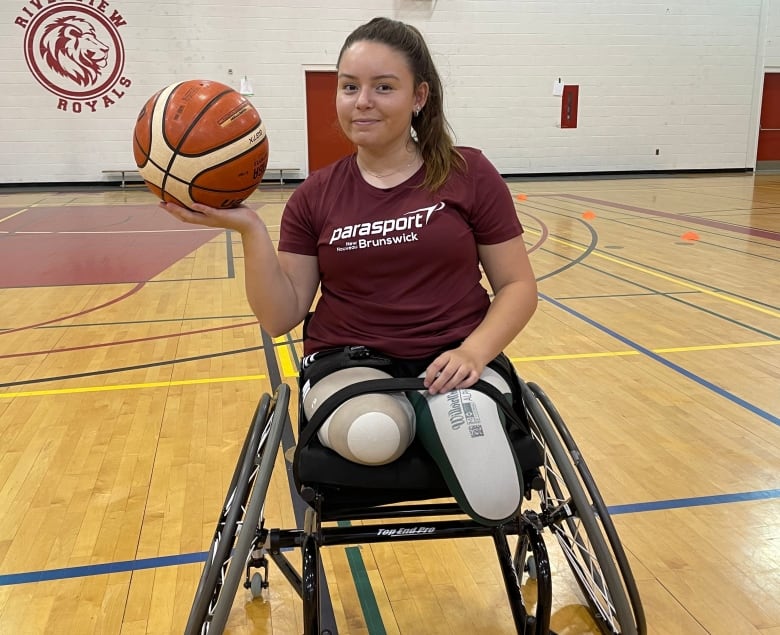 A woman in a wheelchair with brown hair in a ponytail holds up a basketball