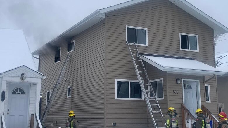 A beige siding building has ladders running up to the windows, firefighters standing at the bottom and smoke coming from the second-storey. 