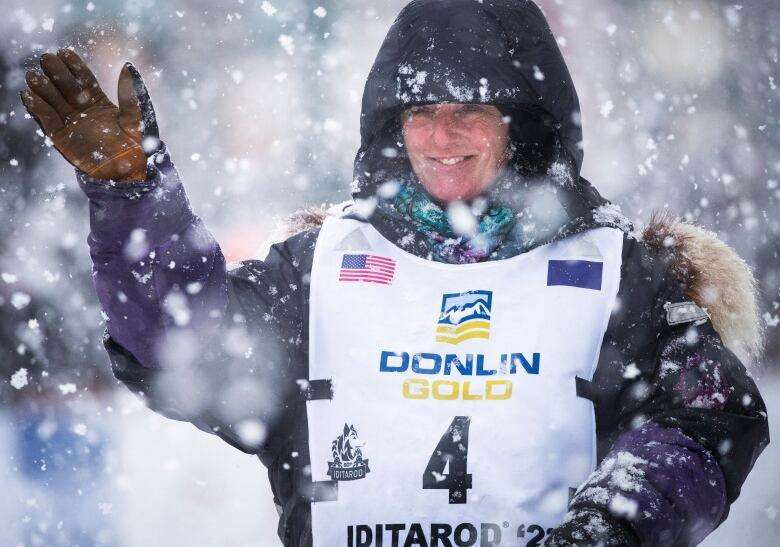 A woman wearing a race bib, mitts and a heavy hooded jacket, waves towards a crowd in the snow, 