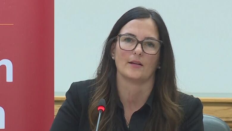 A white woman with long brown hair is wearing glasses and an all-black suit. She is sitting in front of a microphone.