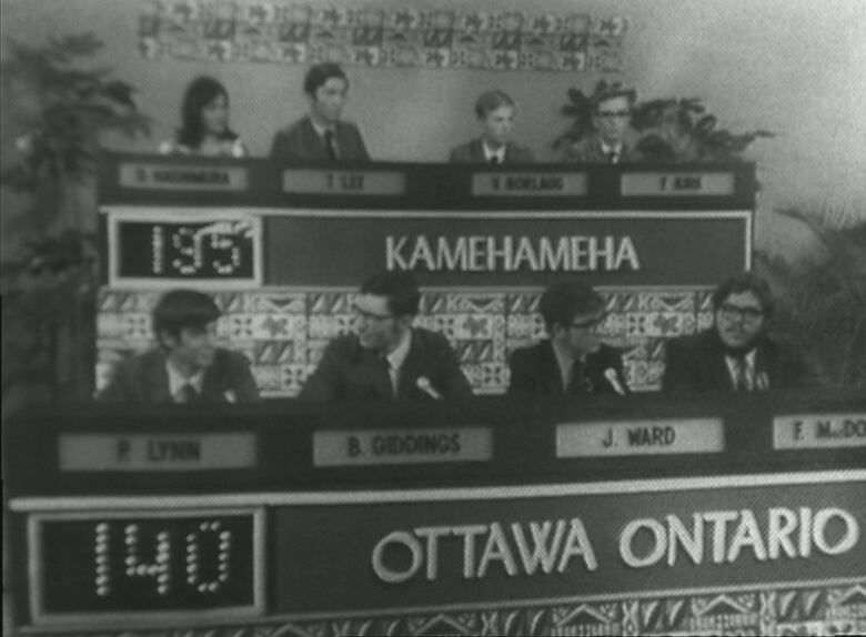 a BLACK AND WHITE PHOTO OF A GAME SHOW WITH four contestants on top of a console and four on bottom with an electronic scoreboard. 