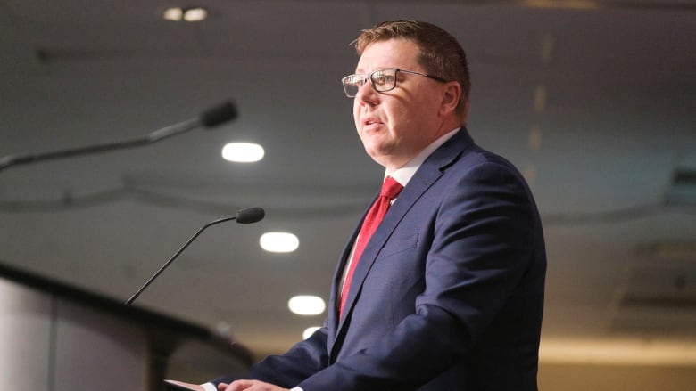 An image of Saskatchewan Premier Scott Moe, wearing a blue suit with a red tie, speaking at a podium with a mic in a room with low lighting.