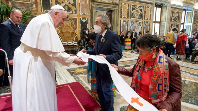 A long, white leather garment that is beaded with orange crosses is pictured being gifted to the Pope by two people.