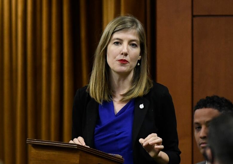 Woman with dirty blonde hair, wearing a bright blue shirt and a black blazer standing behind a podium. 