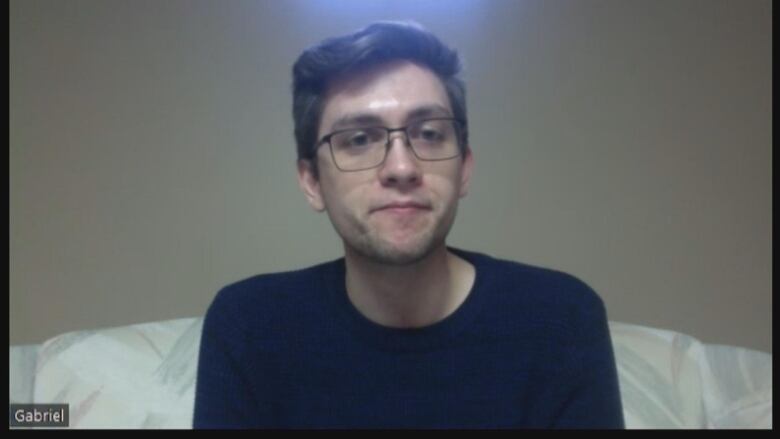 A man with brown hair wearing a blue shirt and glasses sitting on a bed