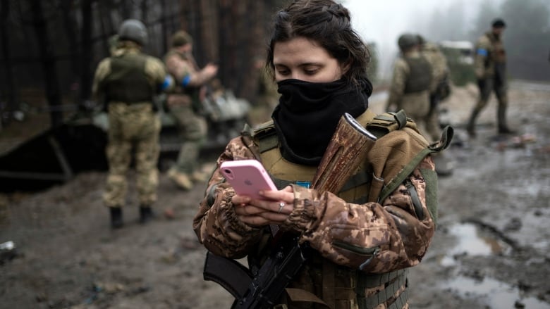 A female solider in full combat gear, standing in a muddy clearing, checks her cellphone, which is pink. 