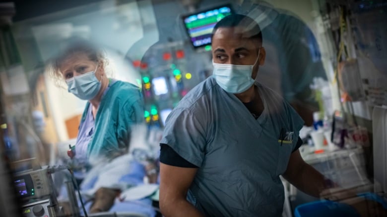 A nurse in a blue medical mask looks away from the camera in a hospital room.