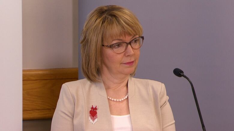 A woman with dyed blonde hair wearing a beige coloured jacket sitting at a microphone.