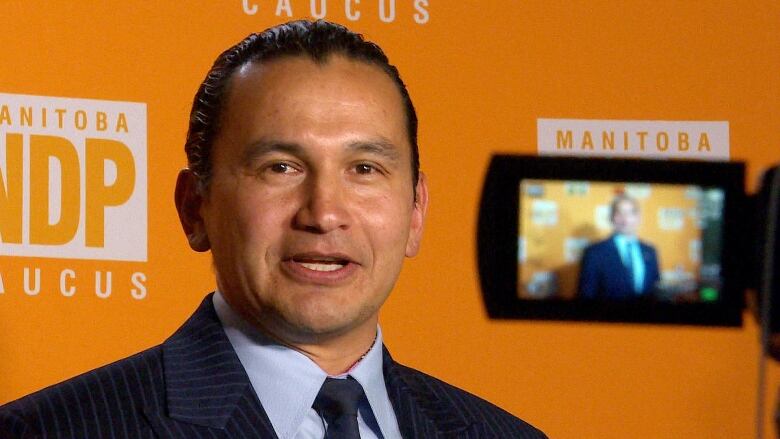 A man with long hair pulled back wears a suit and stands in front of an orange background as he speaks into a camera.