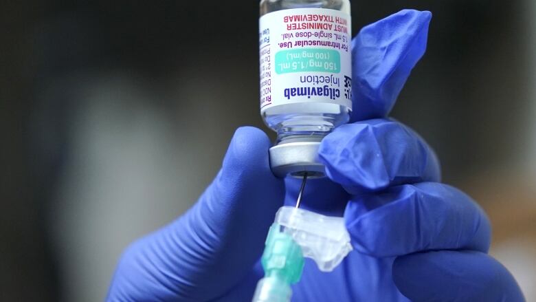 A hand in a blue latex glove holds a small upside-down vial and inserts a syringe into it.