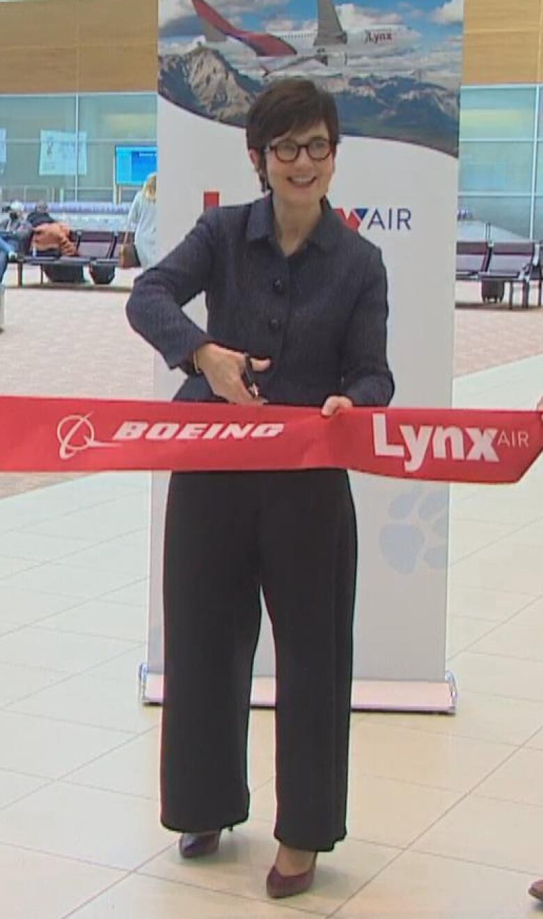 A woman in a dark pantsuit cuts a red ribbon in an airport.