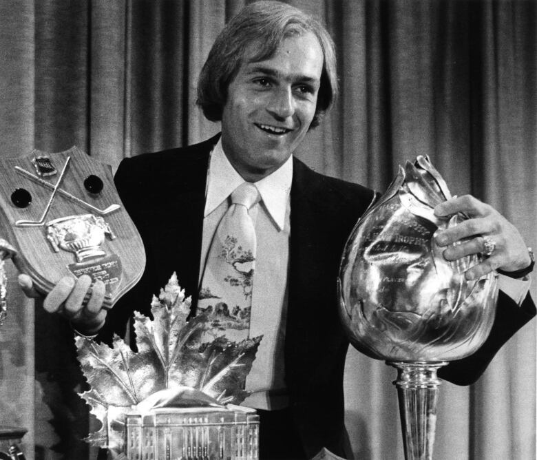 Guy Lafleur faces the camera smiling and holding several trophies. 