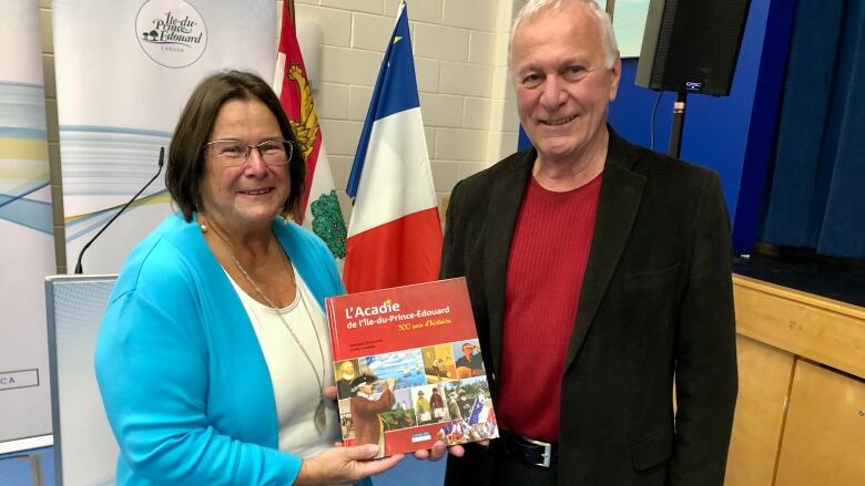 A woman and a man hold a book while standing in front of an Acadian flag.