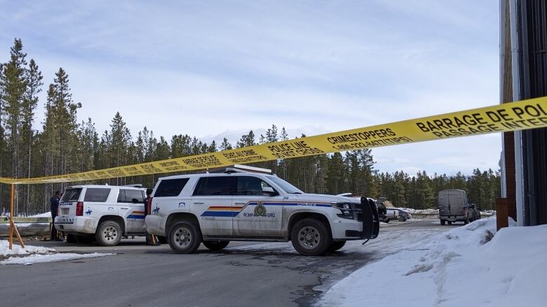 RCMP vehicles are seen behind yellow police tape on a snowy, tree-lined road.