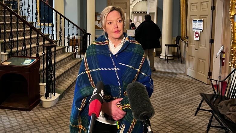 A woman wearing a Nova Scotia tartan poncho stands behind three microphones.