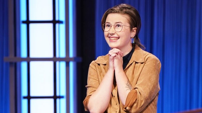 A young woman wearing glasses is smiling as she rests her chin on her folded hands.