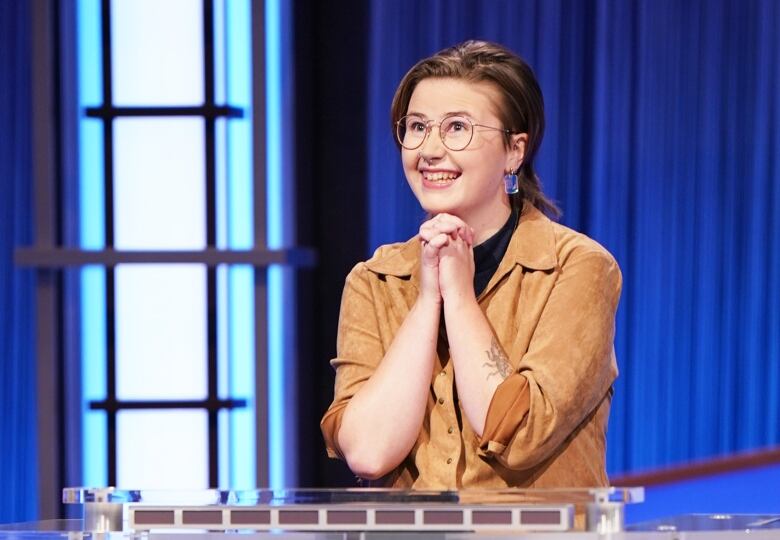 A young woman wearing glasses is smiling as she rests her chin on her folded hands.