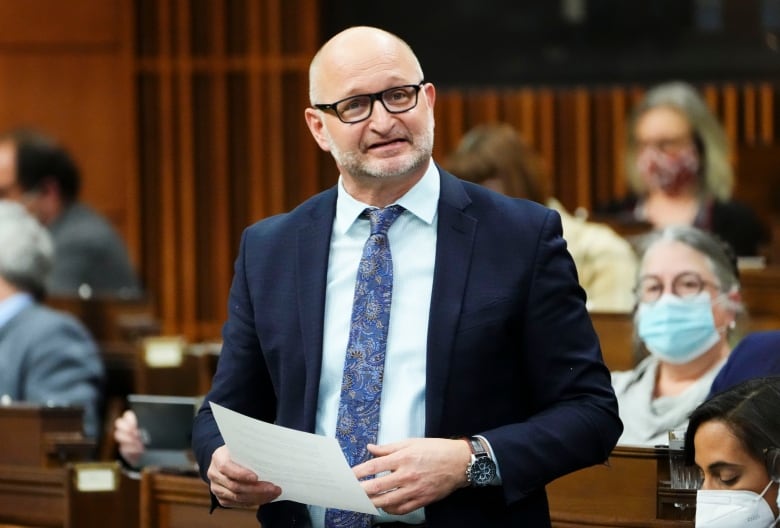 A man in a blue suit, white shirt, and blue tie stands to answer a question in the House of Commons.