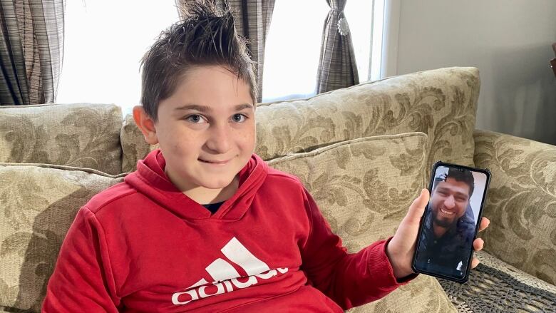 9-year-old boy with red sweatshirt and brown hair holds up cellphone with smiling face of his father.