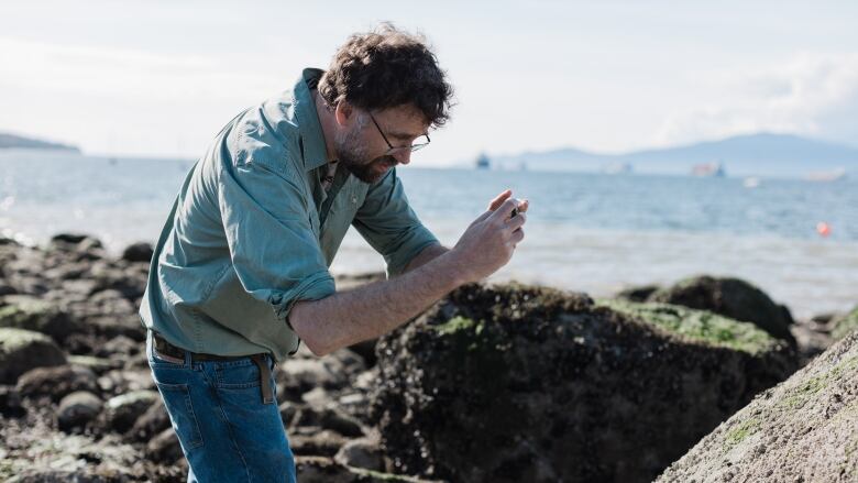 A man in shirt and jeans stoops over a rock to take a picture with a camera.