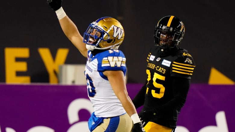 A football player in white and blue with a gold helmet and pants celebrates while another player in black and yellow stands by.