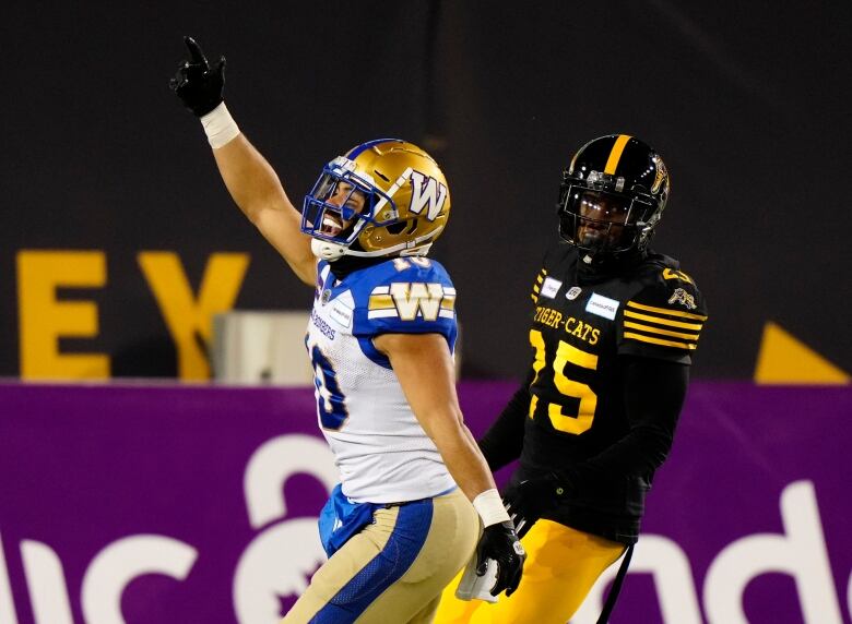 A football player in white and blue with a gold helmet and pants celebrates while another player in black and yellow stands by.