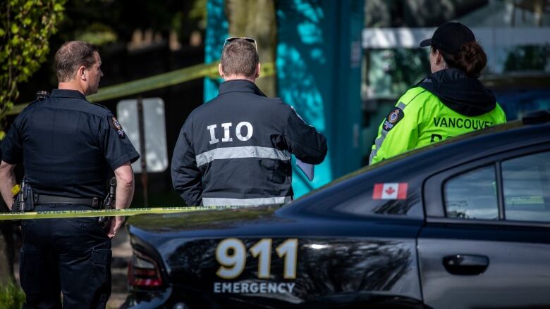 Vancouver police officers and detectives with the Independent Investigations Office stand together wearing branded jackets behind yellow police tape.