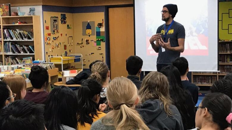 A bearded man in glasses, a winter tuque and Spider-Man t-shirt speaks to students inside a school library. 