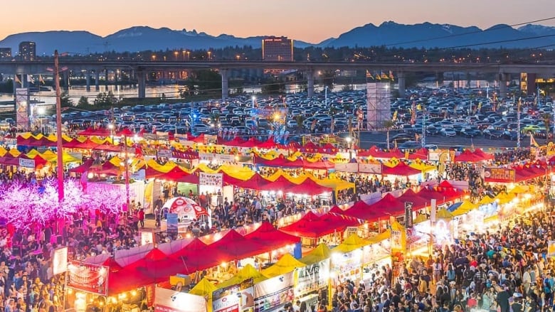 Red and yellow stalls are lined up with thousands of people milling around. In the background, you can see a bridge, the mountains and a pink sunset. 