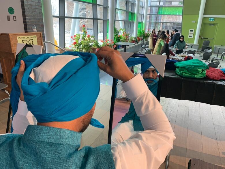 A man looks into a mirror as he ties a blue turban on his head.