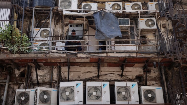 Several air conditioners line three levels of an apartment building and a man sits on a makeshift balcony.