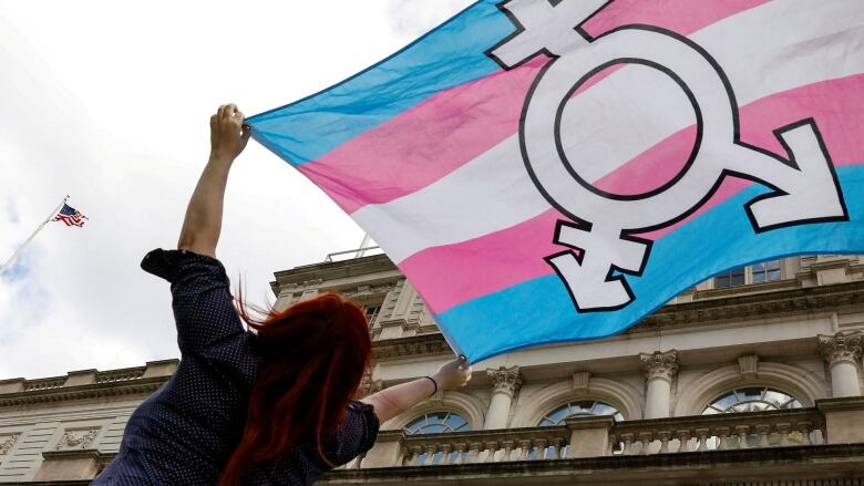 A person with long, bright red hair holds up the pink, blue and white flag of the transgender movement.