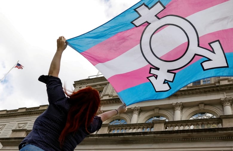 A person with long, bright red hair holds up the pink, blue and white flag of the transgender movement.