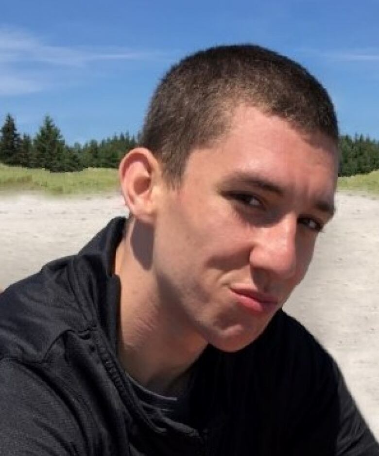 A man with a buzzcut poses for a photo while sitting down on a sandy beach.